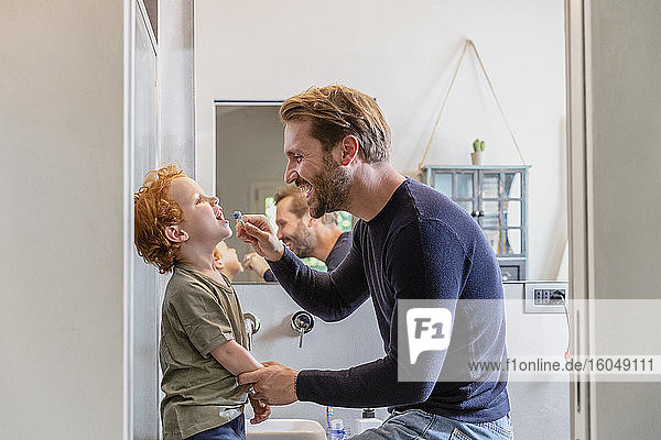 Happy man brushing son's teeth with toothbrush in bathroom at home
