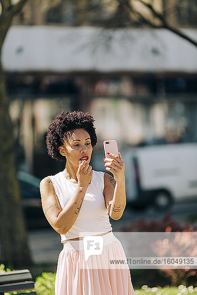 Mid adult woman applying lipstick while looking in smart phone