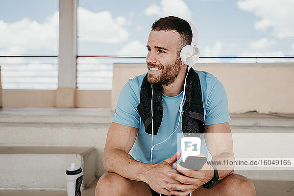 Männlicher Sportler mit Kopfhörern und Smartphone macht eine Pause auf der Tribüne im Stadion