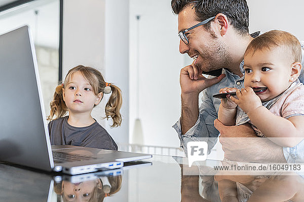 Mann hält Tochter im Gespräch über Smartphone  während Mädchen auf Laptop auf dem Tisch schaut