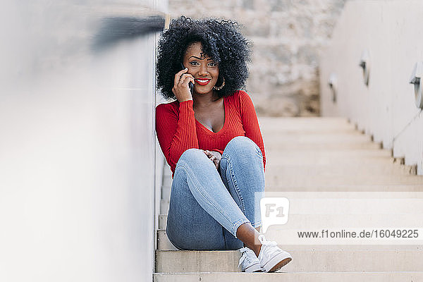 Lächelnde Frau mit Afro-Haar  die auf einer Treppe sitzend ein Smartphone benutzt