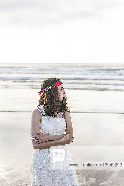 Nachdenkliche junge Frau in weißem Kleid mit verschränkten Armen steht am Meer
