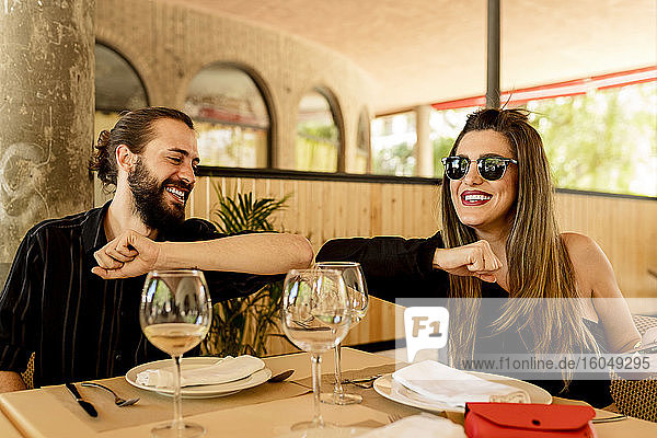 Glücklicher junger Mann und Frau stoßen sich mit dem Ellbogen an  während sie am Tisch im Restaurant sitzen