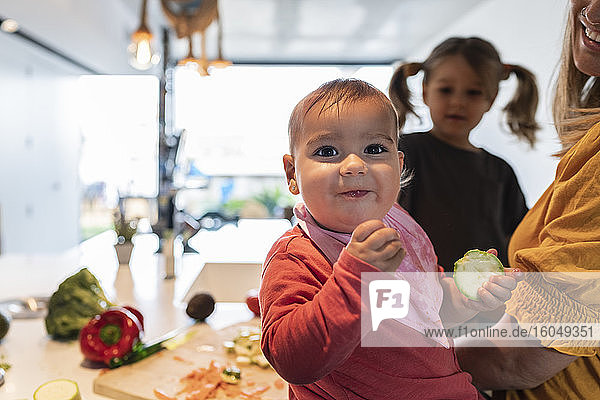Nahaufnahme eines süßen kleinen Mädchens  das Zucchini isst  während Mutter und Tochter im Hintergrund arbeiten