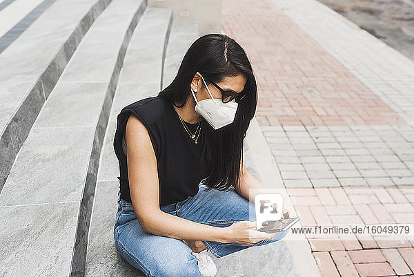 Frau mit Schutzmaske benutzt Smartphone auf einer Treppe in der Stadt