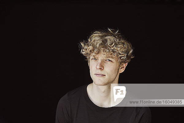 Porträt eines skeptischen jungen Mannes mit lockigem blondem Haar vor schwarzem Hintergrund