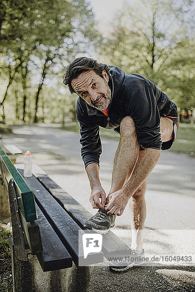 Smiling mature man tying shoelace on bench while standing in park