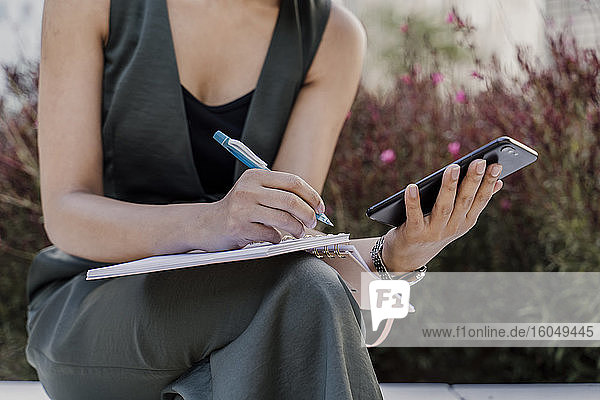 Nahaufnahme einer Geschäftsfrau  die ein Smartphone hält und in einen Notizblock schreibt  während sie im Freien sitzt