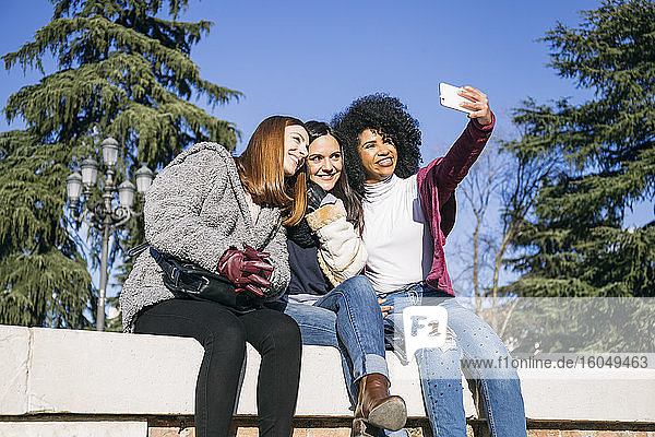 Woman taking selfie with happy female friends while sitting on retaining wall in park
