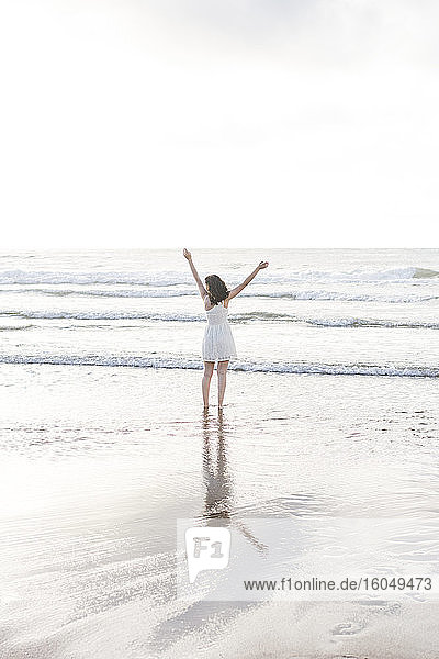 Young woman with arms raised standing at shore against sky