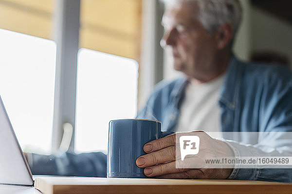 Älterer Mann hält Kaffeetasse  während er zu Hause einen Laptop benutzt