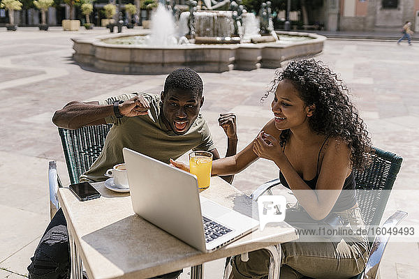 Glückliches Paar gestikuliert während eines Videogesprächs am Laptop in einem Straßencafé
