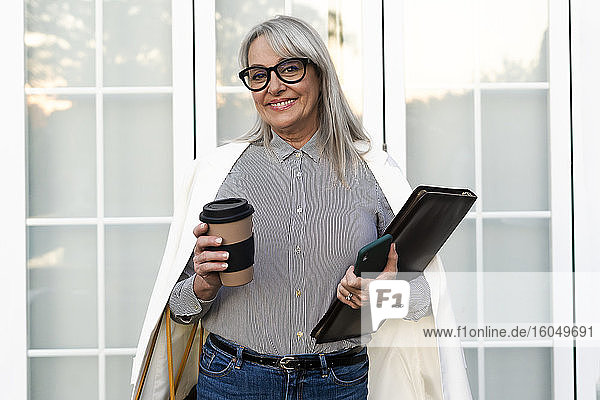 Lächelnde ältere Geschäftsfrau hält Einwegbecher mit Akte und Smartphone vor der Bürotür