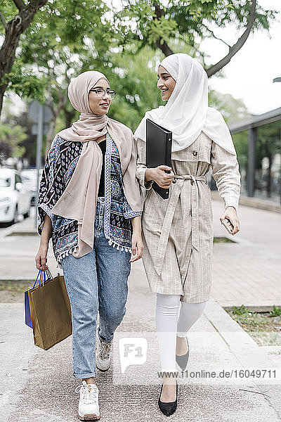 Muslimische Schwestern mit Mappe und Einkaufstaschen unterhalten sich beim Gehen auf dem Bürgersteig in der Stadt