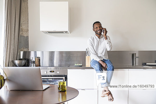 Porträt eines lächelnden Mannes am Telefon  der zu Hause auf dem Küchentisch sitzt