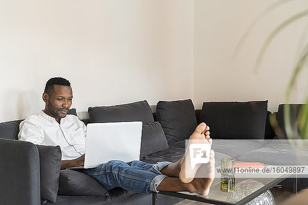 Porträt eines Mannes  der mit hochgelegten Füßen auf der Couch sitzt und einen Laptop benutzt