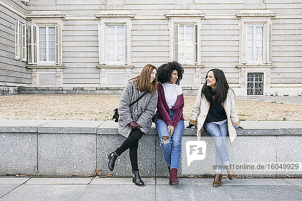 Happy female friends talking while sitting on retaining wall against Madrid Royal Palace  Spain