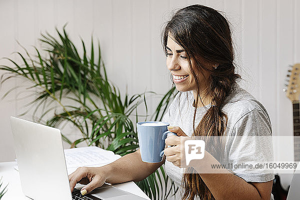 Glückliche junge Frau  die einen Kaffeebecher hält  während sie zu Hause einen Laptop benutzt