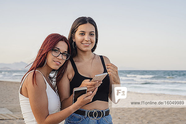 Teenage girls holding smart phones at beach against clear sky