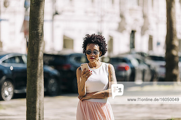 Stilvolle Frau mit lockigem Haar  die in der Stadt stehend ihr Smartphone benutzt