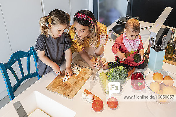 Baby girl sitting on island while mother looking at daughter chopping vegetables in kitchen