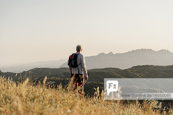 Aktiver älterer Mann mit Rucksack  der bei Sonnenuntergang auf eine Bergkette blickt