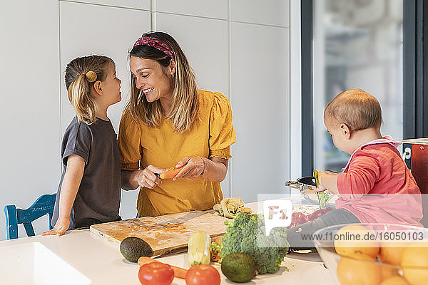 Lächelnde Mutter und Mädchen bereiten Essen zu  während ihre kleine Tochter auf der Kücheninsel spielt