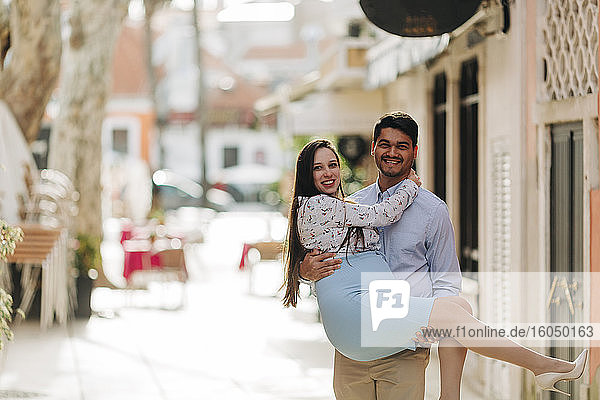 Happy man carrying pregnant woman in city during sunny day