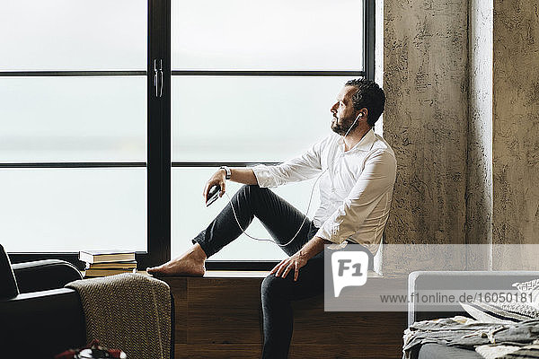 Mature man sitting barefoot on window sill  using smartphone and earphones