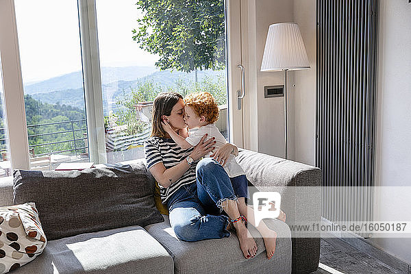 Mother kissing cute son while sitting on sofa in living room at home
