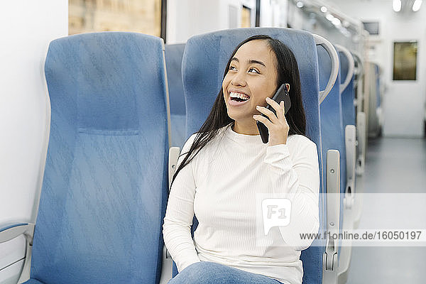 Fröhliche junge Frau  die über ihr Smartphone spricht  während sie auf einem Sitz im Zug sitzt