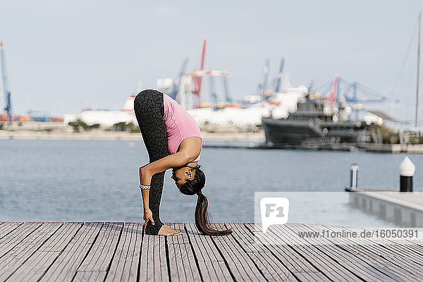 Weibliche Athletin beim Üben der stehenden Vorwärtsbeuge auf der Seebrücke im Hafen