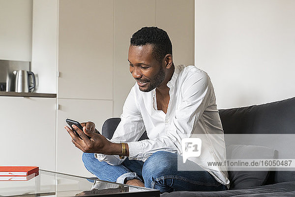 Lächelnder Mann sitzt zu Hause auf der Couch und benutzt sein Smartphone