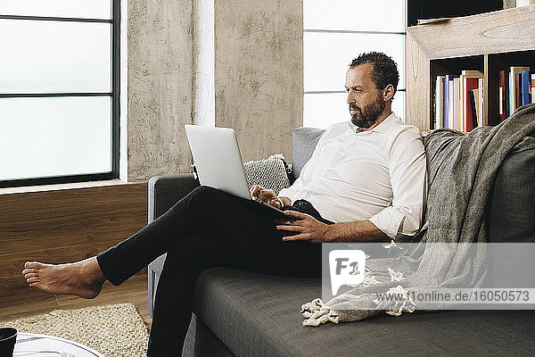 Älterer Mann sitzt auf der Couch und benutzt einen Laptop