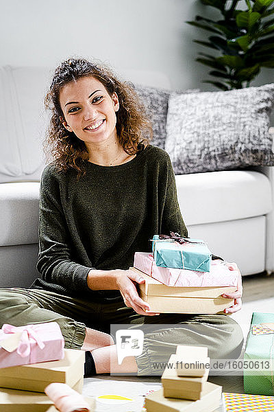Porträt einer lächelnden jungen Frau  die zu Hause mit eingepackten Geschenken auf dem Boden sitzt