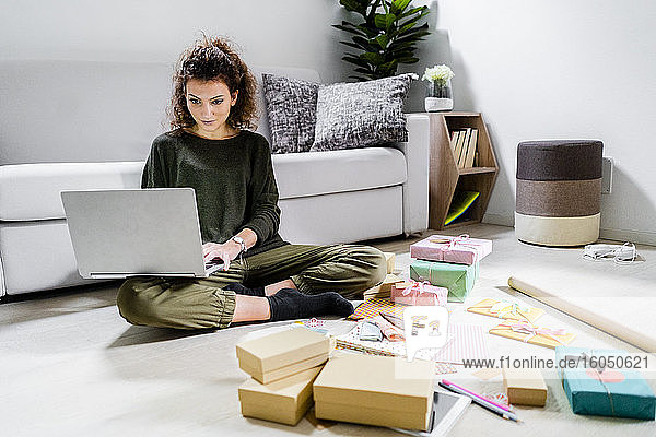 Porträt einer jungen Frau  die mit eingepackten Geschenken auf dem Boden sitzt und einen Laptop benutzt