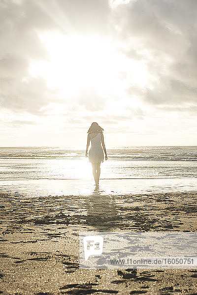 Junge Frau spaziert am Strand gegen den bewölkten Himmel bei Sonnenuntergang