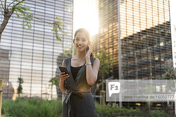 Lächelnde Geschäftsfrau  die ihr Smartphone hält  während sie vor modernen Gebäuden in der Stadt steht