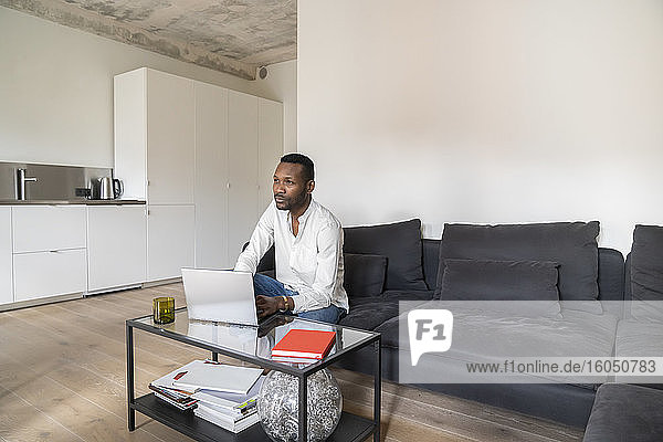 Nachdenklicher Mann sitzt auf einer Couch in einer modernen Wohnung und benutzt einen Laptop