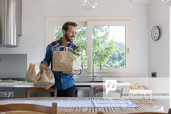 Lächelnder gutaussehender Mann  der eine Tüte mit Lebensmitteln hält  während er in der Küche zu Hause wegschaut