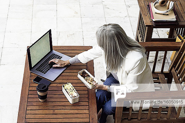 Geschäftsfrau mit grauem Haar  die einen Laptop benutzt  während sie im Garten zu Mittag isst