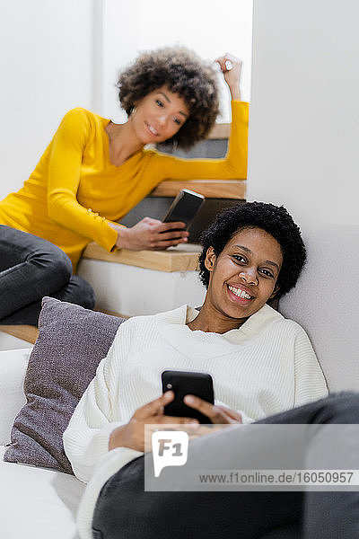 Porträt einer glücklichen jungen Frau  die sich mit ihrem Smartphone auf der Couch entspannt  während ihr Freund im Hintergrund steht