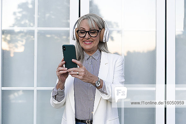 Cheerful businesswoman using smart phone while listening music through headphones against office door
