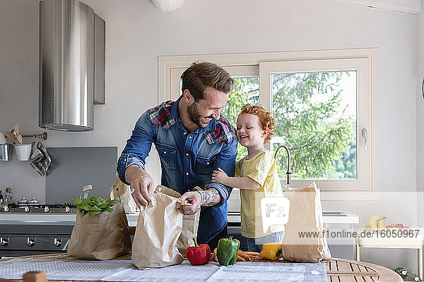 Fröhlicher Vater und Sohn mit Lebensmitteln am Esstisch in der Küche stehend
