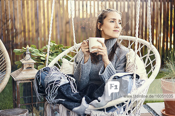 Nachdenkliche junge Frau hält Tasse  während sie sich auf einer Schaukel im Garten entspannt