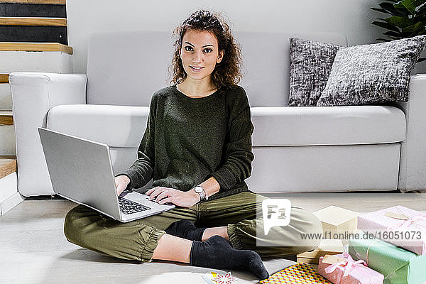 Porträt einer lächelnden jungen Frau  die mit eingepackten Geschenken auf dem Boden sitzt und einen Laptop benutzt