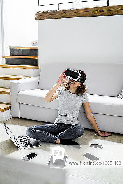 Junge Frau sitzt zu Hause auf dem Boden und benutzt eine Virtual-Reality-Brille und verschiedene elektronische Geräte