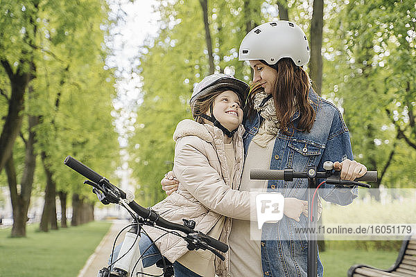 Girl with bicycle embracing mother while standing against trees in city park