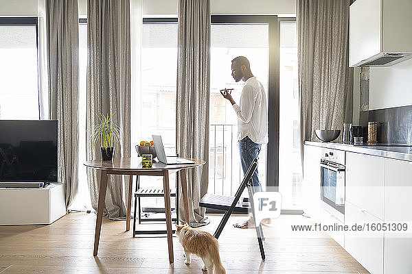 Mann  der in einer modernen Wohnung vor der Balkontür steht und ein Smartphone benutzt