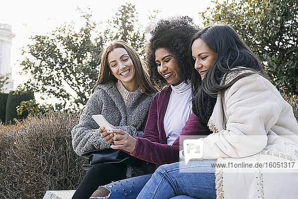 Lächelnde Frau zeigt ihren Freundinnen ihr Smartphone  während sie im Park sitzt